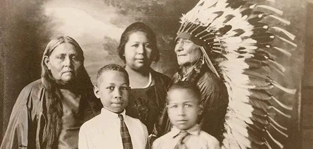A multigenerational Afro-Indigenous family in a formal studio portrait, an elder wearing a feathered headdress