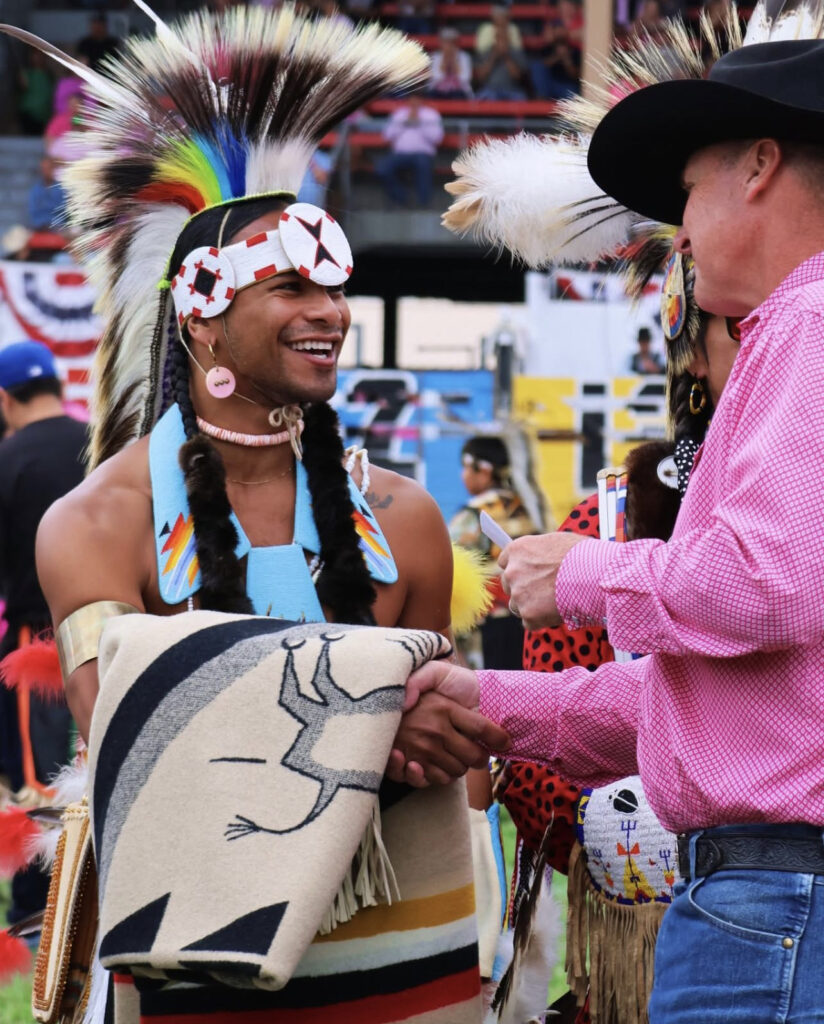 An Afro-Indigenous dancer in full regalia smiling and shaking hands at a powwow