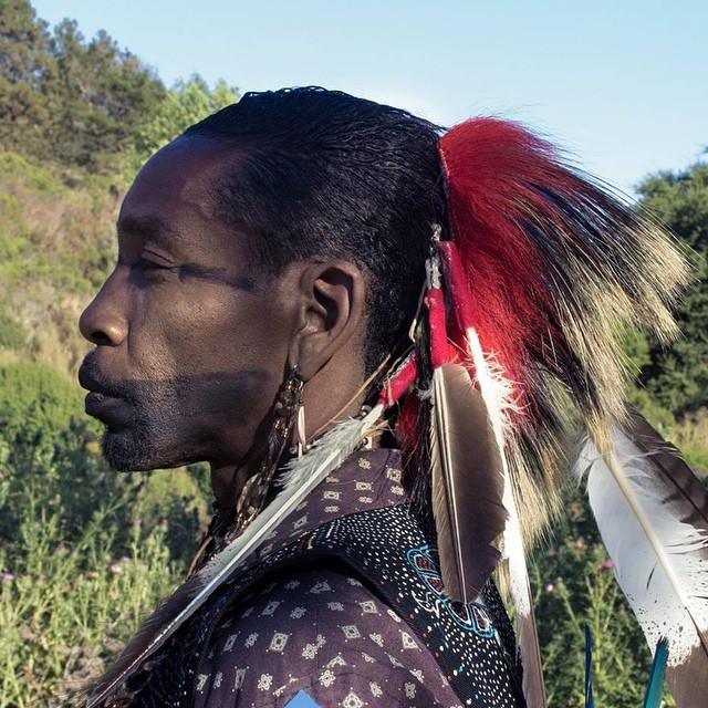 An Afro-Indigenous man in profile wearing feathers and traditional face paint