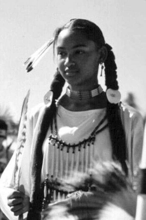 A young Afro-Indigenous woman in traditional regalia with braids, feathers, and beadwork